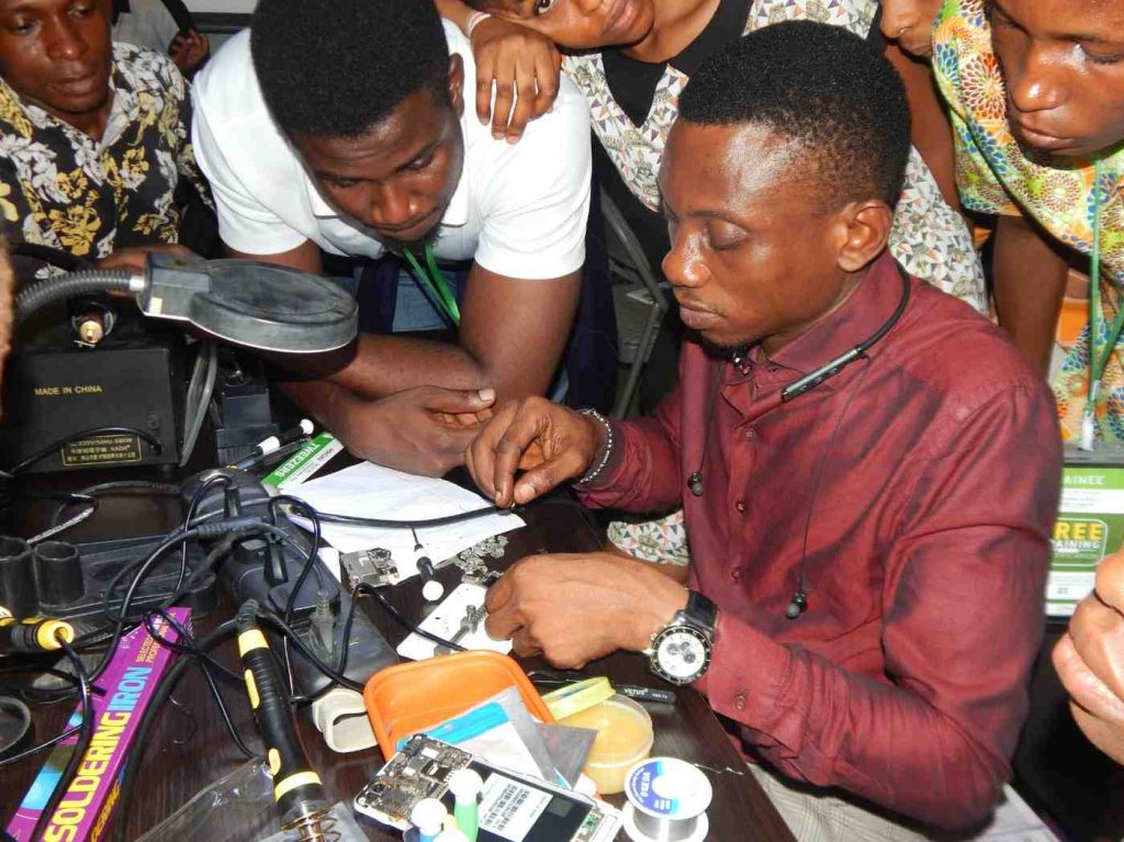 Chidi, an African youth entrepreneur, teaches apprentices to repair phones in a busy Nigerian market stall, tools and devices scattered around, with customers waiting in the vibrant background.