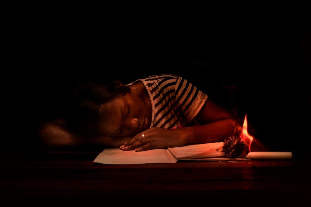 A young woman, one of many African youth entrepreneurs, sketches dress designs by kerosene lamplight in Nigeria, surrounded by fabric swatches, her face glowing with focus and ambition.